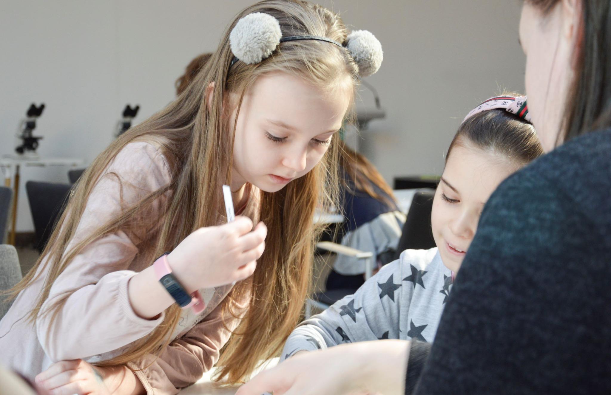 Warsztaty Familijne "Nasz świat pod lupą" - zdjęcia 4 Zdjęcie przedstawia dwie dziewczynki. Jedna z nich ma różową bluzę, a druga szarą w ciemne gwiazdy. Jedna z nich ma w dłoni ołówek, co może oznaczać, że wykonują jakieś zadanie. W tle widać mikroskopy. Zdjęcie: MBP.