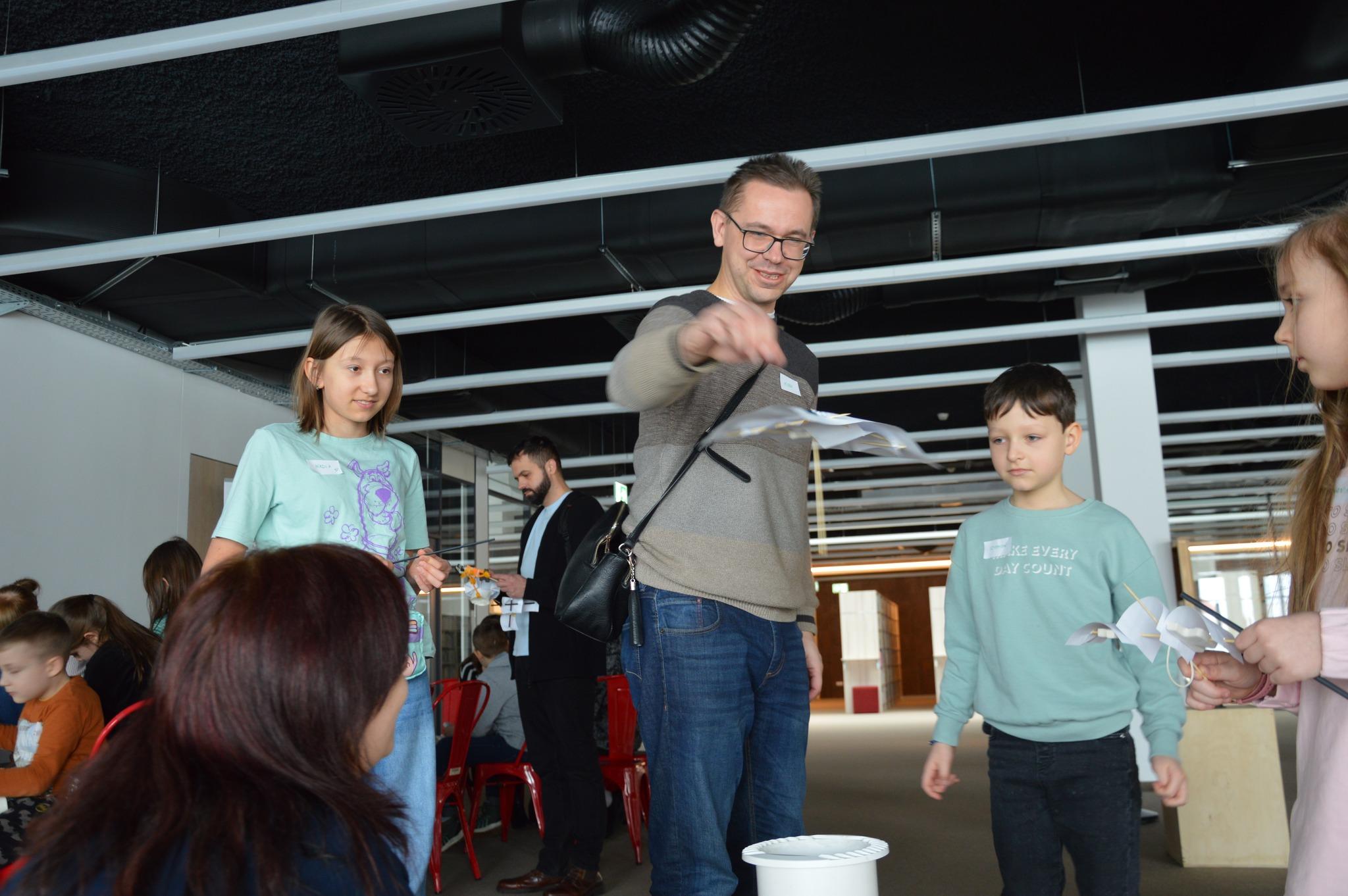 Warsztaty Familijne „Akcja fizyka” - zdjęcia 2 Zdjęcie przedstawia mężczyznę w bluzie utrzymanej w beżowych tonach. Zrzuca on z wysokości konstrukcję zbudowaną z wykałaczki oraz kilku niewielkich kartek. wydaje się być zadowolony z wykonywanej czynności. Wokół mężczyzny stoi trójka dzieci przyglądająca się. W tle widać inne osoby biorące udział w warsztatach. Zdjęcie: MBP.