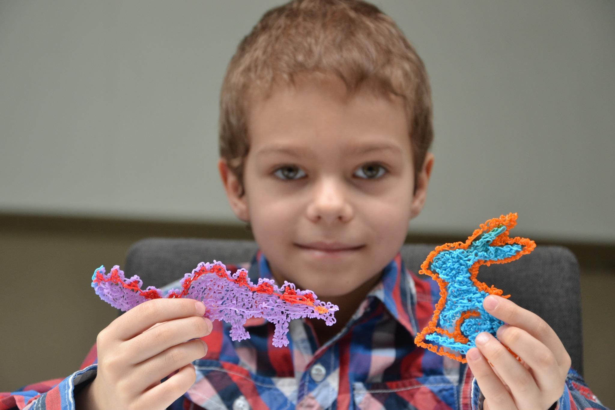 Warsztaty Familijne "Przyjaźń w trzech wymiarach" - zdjęcia 3 Zdjęcie przedstawia chłopca w koszuli w kolorową kratę. Patrzy w obiektyw aparatu i się uśmiecha. W dłoniach trzyma figurki wykonane za pomocą długopisu 3D: w prawej niebiesko pomarańczowego królika, a w lewej fioletowo czerwonego dinozaura. Zdjęcie: MBP.