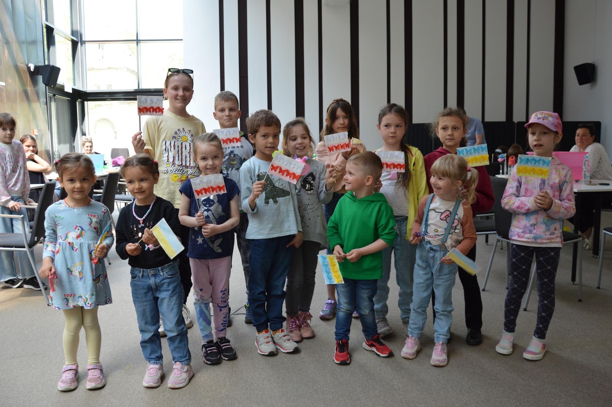 Warsztaty Familijne "Statek Odkrywców" - zdjęcia 12 Zdjęcie przedstawia grupę dzieci. Każde z nich trzyma w dłoniach wykonaną z papieru flagę. Część dzieci ma flagi Polski, część flagi Ukrainy. Na każdej fladze przyklejone są motylki w odpowiadających kolorach. Zdjęcie: MBP.