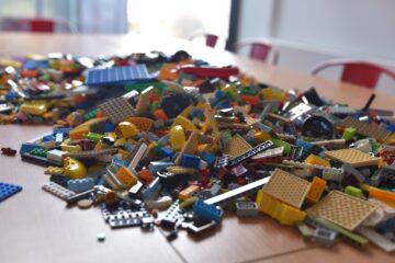 LEGO w Mediatece 2 Na jasnym drewnianym stole leżą rozrzucone klocki Lego w różnych kolorach. W tle widać czerwone metalowe krzesła oraz duże okno. Zdjęcie: MBP.