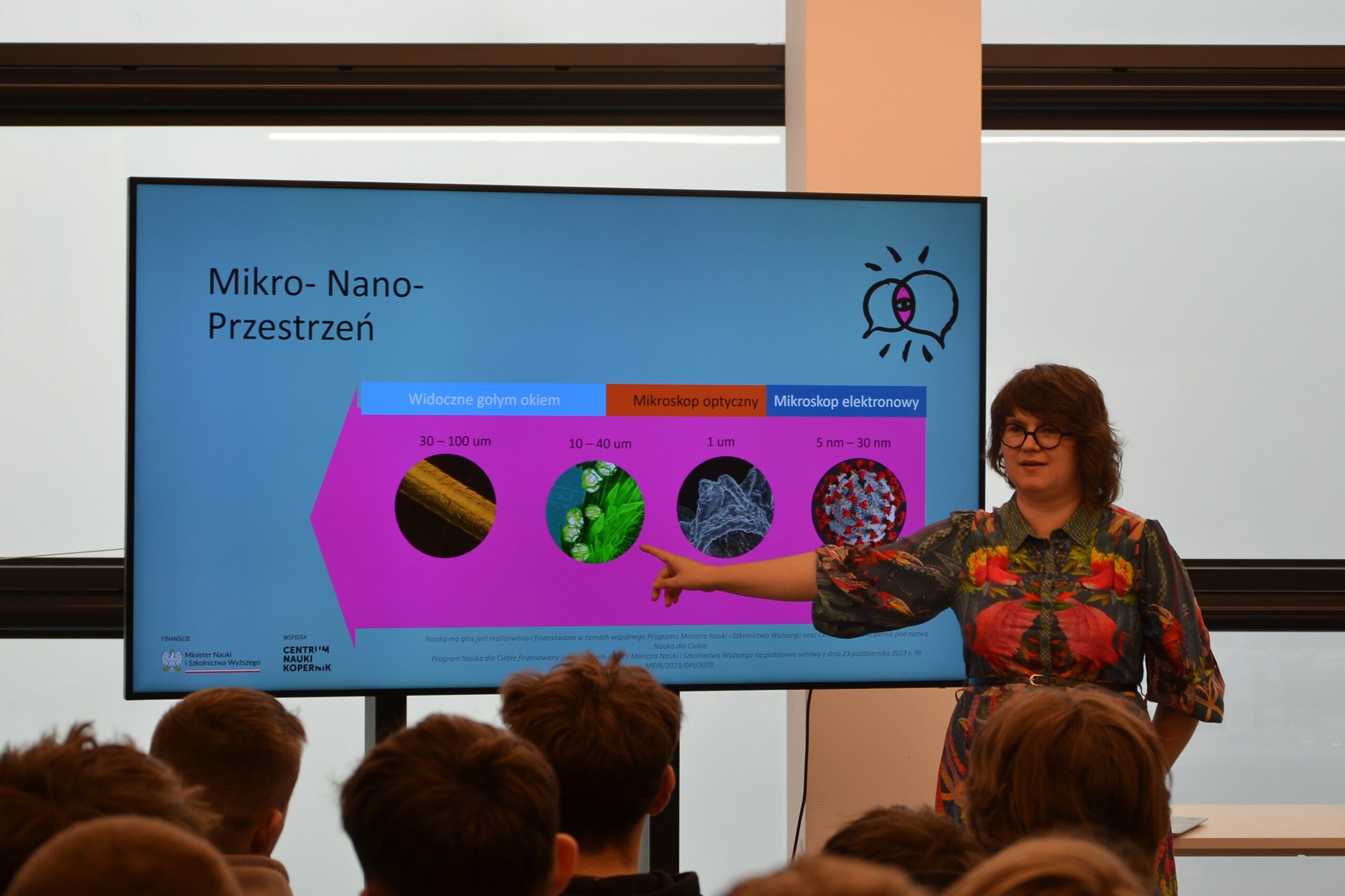 Nauka ma głos - spotkanie 6 Zdjęcie prezentuje prawdopodobnie wykład. W centrum zdjęcia znajduje się stojąca osoba w kolorowej sukience. Kobieta ma krótkie włosy i okulary. Wyciągniętą ręką pokazuje na grafikę znajdującą się na monitorze. Obok niej widoczne są jeszcze trzy grafiki i opis. Prezentacja jest w kolorach niebieskim i różowym. Widoczny jest napis Nauka ma głos. Wydarzenie ma miejsce w niewielkim pomieszczeniu. Widać od tyłu siedzące młode osoby. Zdjęcie własnością MBP.