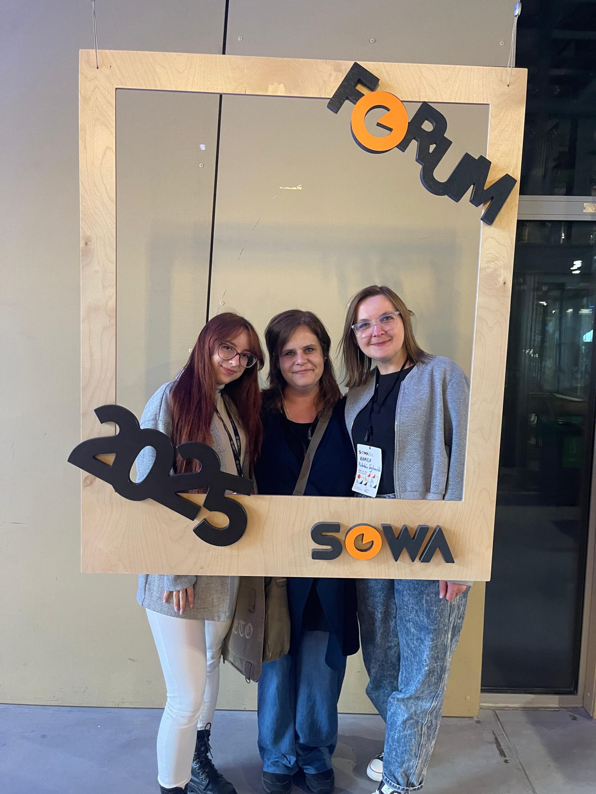 Forum SOWA 2025 8 Zdjęcie przedstawia trzy kobiety. Dwie stojące z zewnątrz mają okulary. Osoba stojąca z lewej ma włosy w kolorze czerwonym. Po środku stoi szatynka, a z prawej strony blondynka. Przed osobami znajduje się zawieszona drewniana ramka. W górnym prawym rogu jest skośny napis Forum. W lewym dolnym rogu znajduje się rok 2025. W prawym dolnym rogu ramki jest napis SOWA. W tle widać pastelową ścianę oraz przeszkloną przestrzeń. Mogą to być drzwi. Zdjęcie zrobione zostało w Centrum Nauki Kopernik. Zdjęcie własnością MBP.
