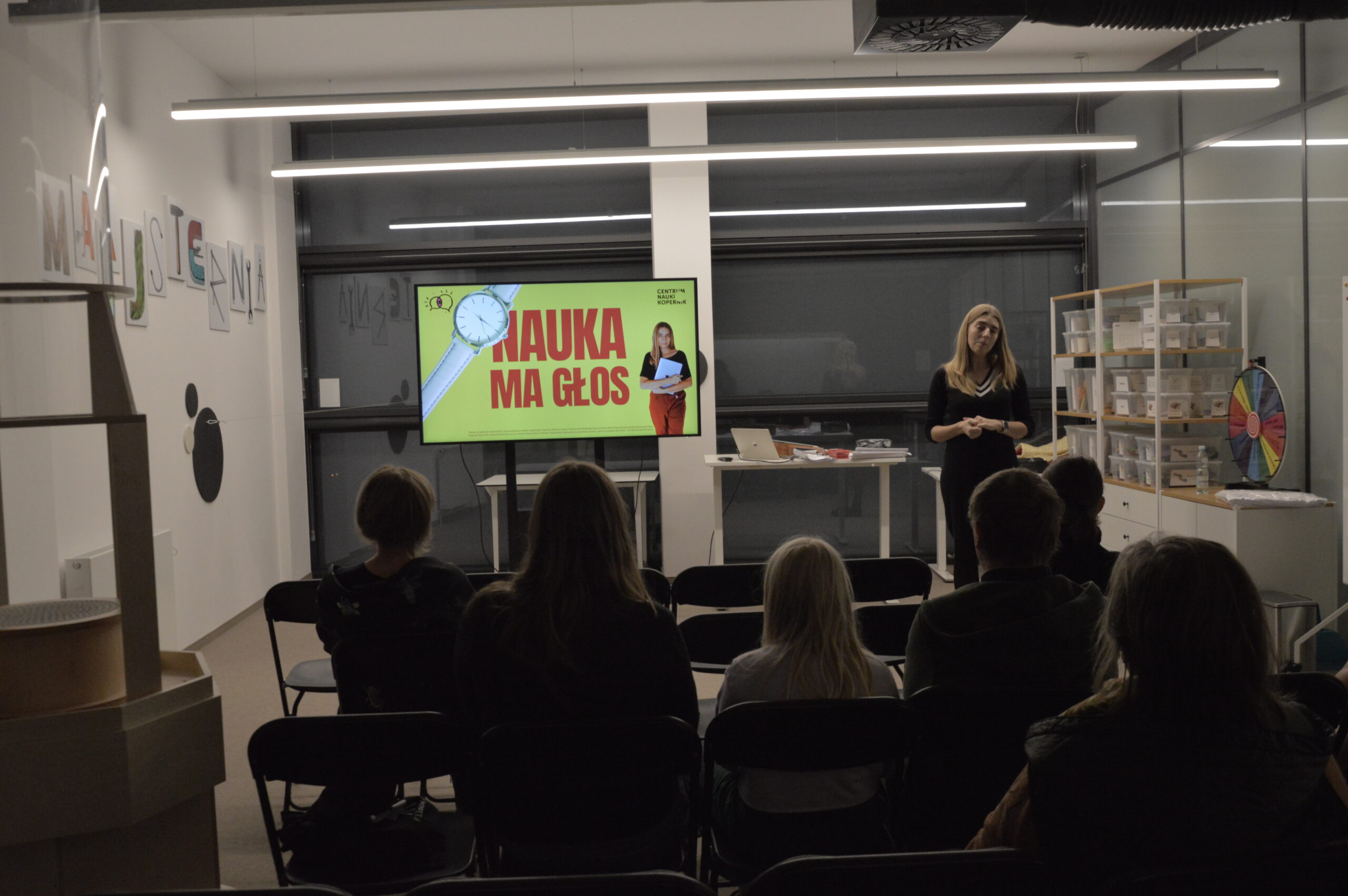 Zdjęcie przedstawiające spotkanie z kobietą w czarnym stroju. Obok niej stoi duży ekran, na którym wyświetlona jest grafika: na żółtym tle widać czerwony napis "Nauka ma głos". Na literze N leży biały zegarek na rękę z tarczą. Po prawej stronie grafiki widać zdjęcie kobiety prowadzącej spotkanie. Obok ekranu widać stół, na którym znajduje się laptop oraz inne elementy. Po prawej stronie zdjęcia widać dużą półkę, na której stoją plastikowe pudełka. Po lewej stronie, na ścianie wisi napis "Majsternia", wykonany z grafik przedstawiających narzędzia. Zdjęcie: MBP.